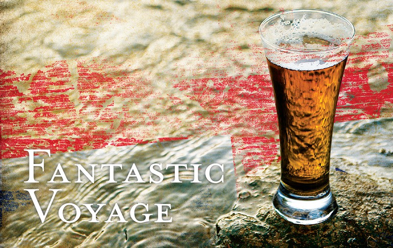 A tall glass of IPA sits nestled on an oceanside rock as small wavelets lap the shore.