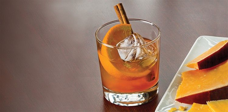 Rum old fashioned garnished with an orange wedge and cinnamon stick with a plate of aged Gouda on the side