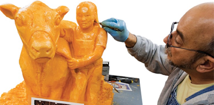 man carving a piece of cheese into a likeness of a girl and her prize-winning cow