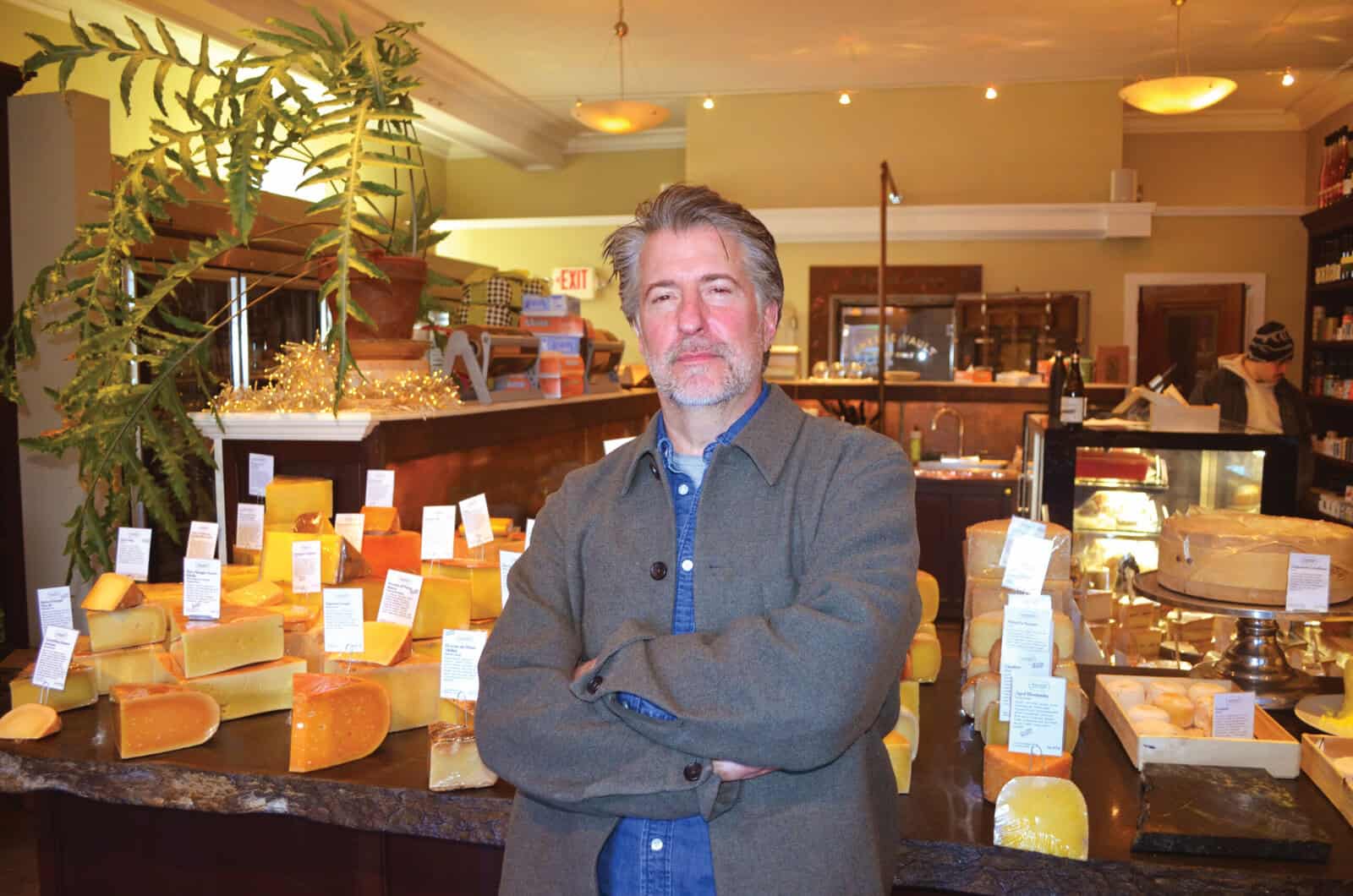 Man stands with arms folded with cheese display behind.